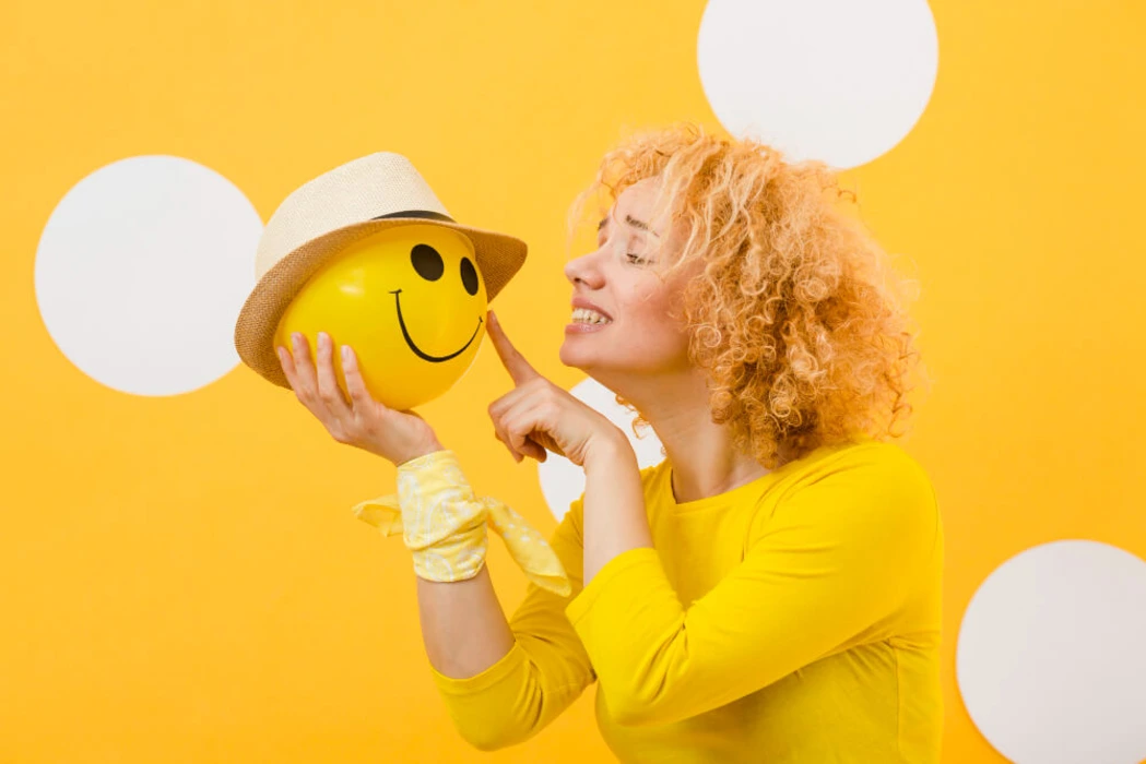 A girl holding a yellow color scheme emoji and similing at it while holding the yellow color emoji in her hand.